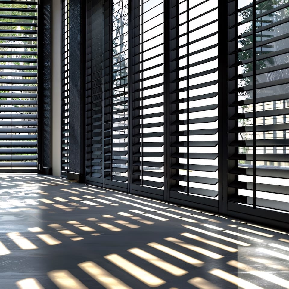 Sunlight streams through closed black shutters in a modern room, casting long, horizontal shadows on the floor.